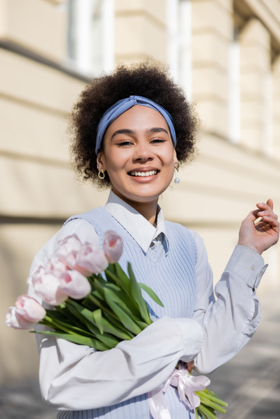 cheerful young african american woman in blue vest and white shirt holding bouquet of tulips - Foto, imagen