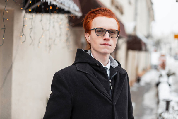 pohledný rusovlasý hipster s brýlemi v módním oblečení s kabátem stojí ve městě a dívá se do kamery. Stylový obchodník venku v zimním dni - Fotografie, Obrázek