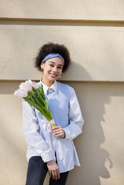 cheerful african american woman in blue vest and white shirt holding bouquet of tulips near wall - Photo, Image