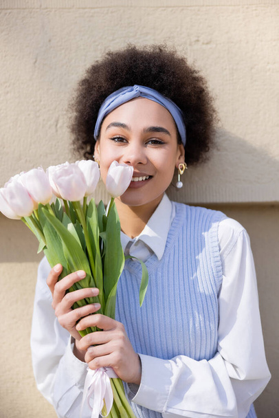 smiling african american woman in blue vest and white shirt holding bouquet of tulips near wall - Fotografie, Obrázek