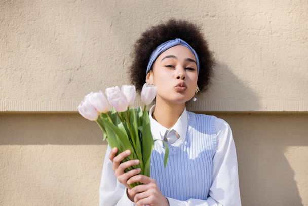 curly african american woman in blue vest and white shirt holding bouquet of tulips and pouting lips near wall - Photo, Image