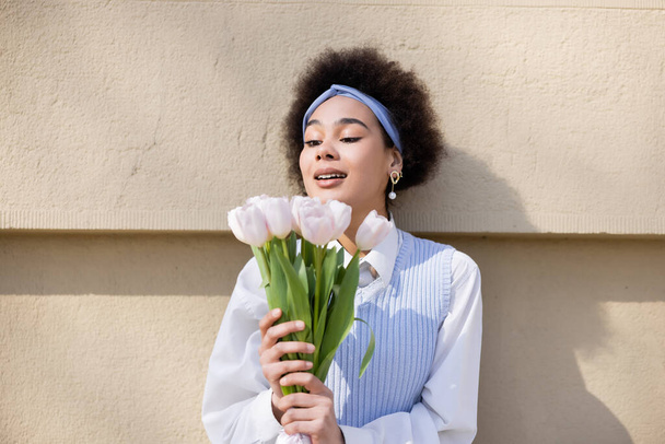 amazed african american woman in blue vest and white shirt holding bouquet of tulips near wall - Photo, Image