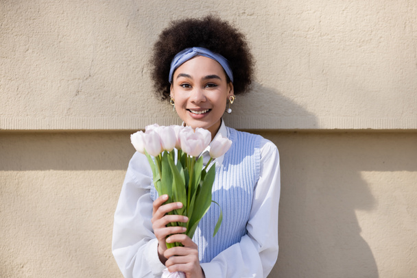 joyful african american woman in blue vest and white shirt holding bouquet of tulips near wall - Photo, Image