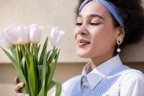 cheerful african american woman in blue vest and white shirt looking at tulips near wall - Foto, Bild
