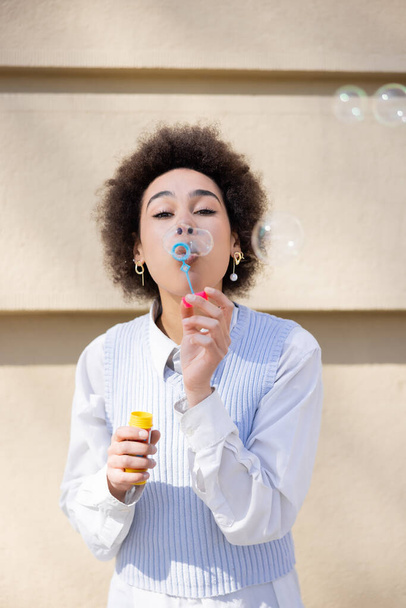young african american woman in blue vest and white shirt blowing soap bubbles - Foto, imagen