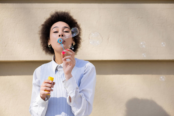 curly african american woman holding bottle and blowing soap bubbles near wall - Valokuva, kuva
