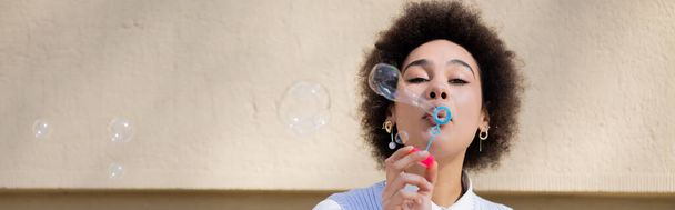 curly african american woman blowing soap bubble near wall, banner - Foto, Imagem