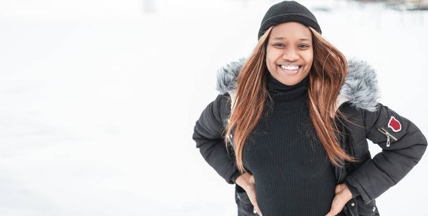 Hermosa joven elegante chica negra con una sonrisa en la ropa de invierno de moda con una chaqueta y sombrero en la nieve blanca - Foto, Imagen