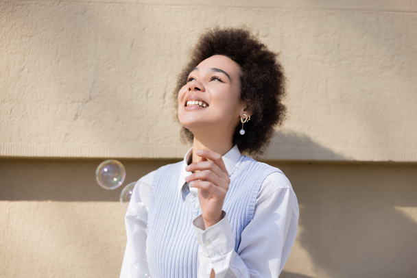 mujer afroamericana positiva sonriendo cerca de burbujas de jabón - Foto, Imagen