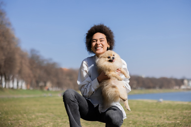 Χαρούμενη Αφροαμερικανή που κρατάει στα χέρια της πομεράνιαν σκύλο. - Φωτογραφία, εικόνα