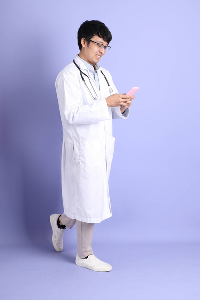 The young adult Asian physician standing on the purple background. - Φωτογραφία, εικόνα