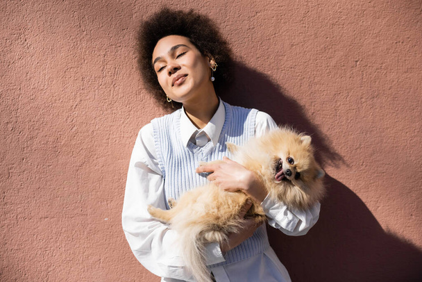 cheerful african american woman in blue vest holding in arms pomeranian spitz near wall - Photo, image