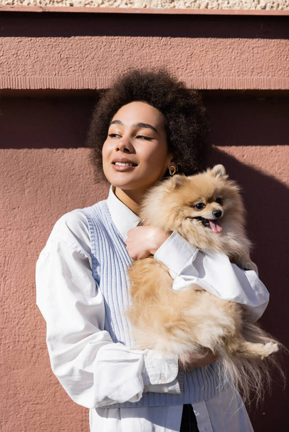 happy african american woman in blue vest holding in arms pomeranian spitz near wall - Valokuva, kuva