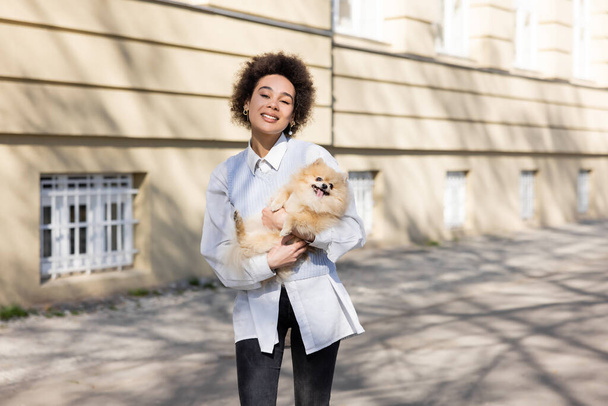 positive african american woman in blue vest holding pomeranian dog on street - Photo, Image