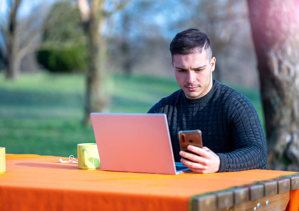 Retrato de atraente jovem trabalhador de escritório em desgaste casual, ele está olhando para sua tela do celular. Eu trabalho remotamente com o laptop de uma mesa no parque. - Foto, Imagem
