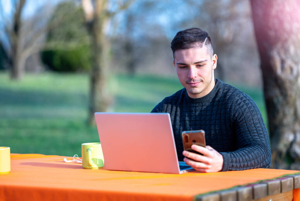 Portret atrakcyjnego młodego pracownika biurowego w luźnym ubraniu, patrzy na ekran telefonu komórkowego. Pracuję zdalnie z laptopem ze stołu w parku.. - Zdjęcie, obraz