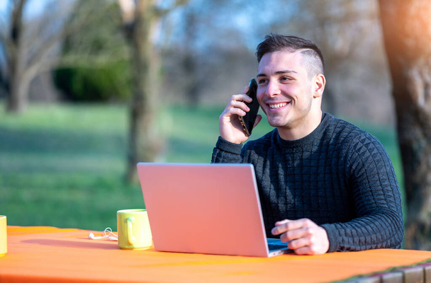 Portrait of attractive young office worker in casual wear, receives a phone call on his cellphone, smiles as he answers. I work remotely with the laptop from a table in the park. - Photo, Image