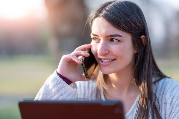 Porträt einer jungen schönen Frau, die fernab in einem Park arbeitet, auf ihrem Handy spricht und lächelt, während sie auf der Tastatur ihres Tablets tippt. Konzept für ferngesteuertes Arbeiten im Freien. - Foto, Bild