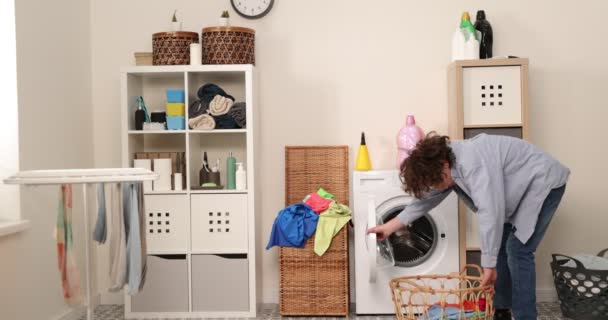 Ein lächelnder Teenie-Junge in Jeans und Hemd sitzt vor einer heimischen Waschmaschine. Er lädt schmutzige Wäsche in die Waschmaschine. Eine helle und luftige Waschküche - Filmmaterial, Video