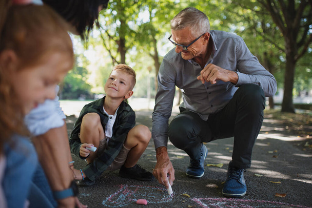 Senior couple with grandchildren drawing with chalks on pavement outdoors in park. - Fotoğraf, Görsel