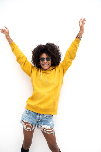 Happy smiling girl with afro hairstyle posing on the white wall, wearing yellow blouse, short jeans and fashionable sunglasses. A lot of copy space. - Photo, image