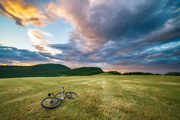 Sunset dramatic sky over meadow in Marche region, Italy. MTB lying on grass amid unique hills and mountains landscape. Traveling by bicycle, alternative mobility concept. - Photo, Image