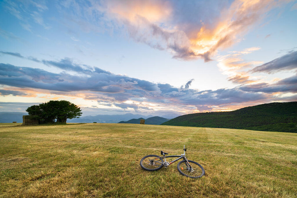 Puesta de sol cielo dramático sobre el prado en la región de Marche, Italia. MTB acostado en la hierba en medio de colinas y montañas únicas paisaje. Viajar en bicicleta, concepto de movilidad alternativa. - Foto, Imagen
