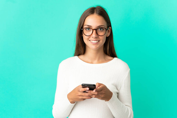 Młoda kobieta na odizolowanym niebieskim tle patrząc w kamerę i uśmiechając się podczas korzystania z telefonu komórkowego - Zdjęcie, obraz