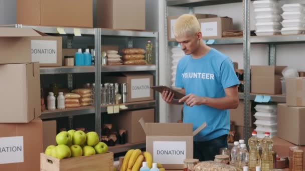 Сосредоточенный волонтер-мужчина использует планшет для упаковки коробки для пожертвований - Кадры, видео