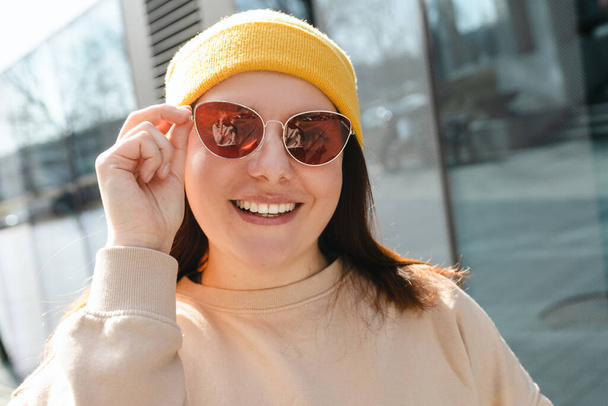 Feliz joven alegre caminando por la calle de la ciudad. Retrato de mujer hermosa en sombrero amarillo y gafas de sol rosadas sonriendo al aire libre. Concepto de estilo de vida urbano. Viajero - Foto, Imagen