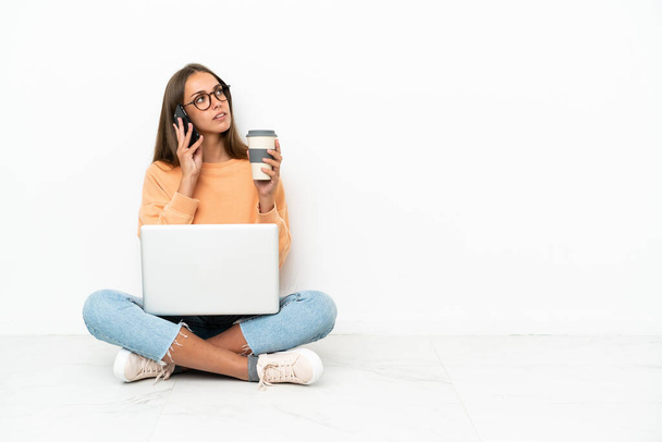 Jeune femme avec un ordinateur portable assis sur le sol tenant du café à emporter et un mobile - Photo, image