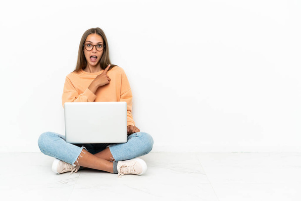 Молодая женщина с ноутбуком, сидящая на полу удивлена и указывая стороны - Фото, изображение