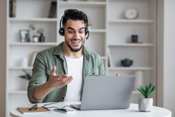 Tutoría en línea. Hombre árabe sonriente en auriculares teniendo videoconferencia en el ordenador portátil - Foto, imagen