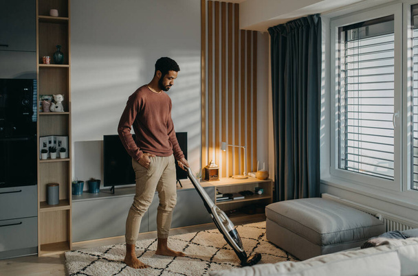 Junger Mann staubt Teppich mit Staubsauger im Wohnzimmer - Foto, Bild