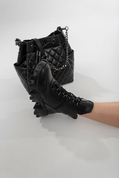 Γυναικείο πόδι με δερμάτινη μοντέρνα μπότα και μαύρη κομψή τσάντα σε λευκό φόντο. Κορίτσι με κομψά παπούτσια και vintage τσάντα. Minimalism έννοια μόδας - Φωτογραφία, εικόνα