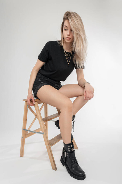 Sexy jolie femme blonde à la mode vêtements noirs avec t-shirt, short et chaussures en cuir est assis sur une chaise en bois. Beauté fille modèle en studio - Photo, image