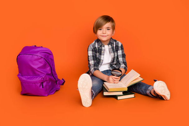 Plná velikost fotografie mladého chlapce sedět na podlaze číst knihu pomocí lupa materiály izolované přes oranžové barvy pozadí - Fotografie, Obrázek
