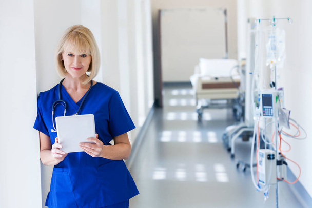 Portrait smiling blonde Caucasian female clinical hospital nurse in scrubs using wireless tablet at modern medical center - Foto, imagen