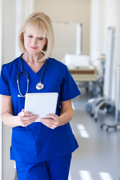 Kavkazská americká žena blondýny senior ošetřovatelství nemocniční personál pomocí bezdrátové dotykové obrazovky tablet v nemocniční chodbě - Fotografie, Obrázek