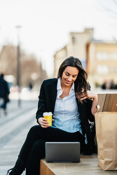 Happy young business woman sitting on street bench and using her laptop computer for work and communication. She is smiling and looking at side. Business on the go and mobile office concept. Bright sunny day. - Foto, Imagen