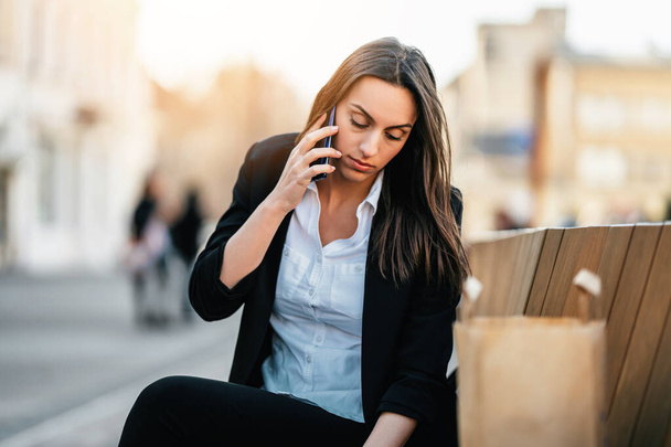Confident young business woman sitting on street bench and using her laptop computer for work and communication. She is serious and looking at side. Business on the go and mobile office concept. Bright sunny day.  - 写真・画像