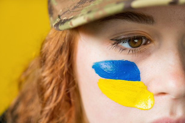 Red-haired woman with tragic look, military hat and national flags of Ukraine on her cheeks and on the background - Fotó, kép
