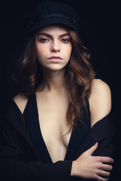 Fashionable studio portrait of a beautiful young model in black hat and sexy top on a black background. Low key. - Photo, Image