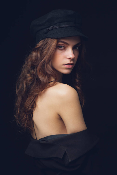 Fashionable studio portrait of a beautiful young model in black hat and sexy top on a black background. Low key. - Fotó, kép
