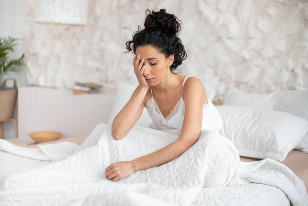 Väsynyt unelias melko tuhatvuotinen eurooppalainen brunette nainen herää yksin töissä, istuu sängyllä valkoisessa makuuhuoneessa - Valokuva, kuva