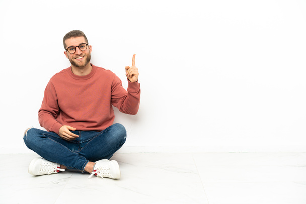 Joven hombre guapo sentado en el suelo mostrando y levantando un dedo en señal de la mejor - Foto, Imagen
