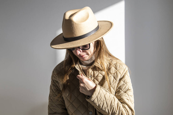 Jovem mulher em uma jaqueta em óculos de sol em um chapéu contra uma parede branca  - Foto, Imagem