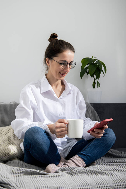Fiatal nő szemüvegben, telefonnal, egy csésze kávéval a kanapén. - Fotó, kép