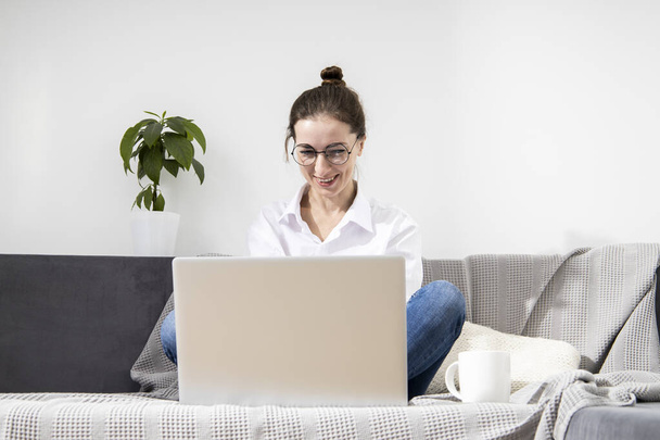Smiling young woman in glasses using laptop sitting on sofa - 写真・画像