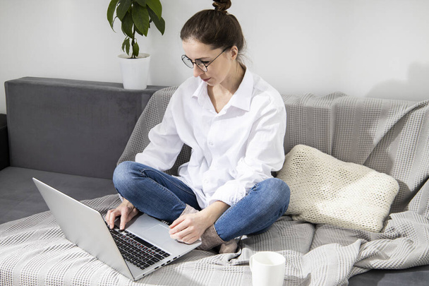 Young woman in glasses with laptop with coffee sitting on sofa - Фото, зображення